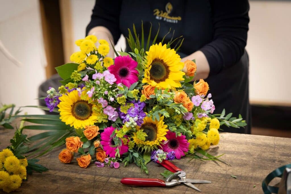 Bright birthday flower bouquet of sunflowers, gerberas, and roses being arranged on a wooden table by a Williamson's florist